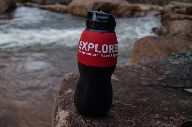 Water bottle with "EXPLORE The Adventure Travel Experts" text, standing upright on a rock near a flowing river. The background features water and surrounding rocks, suggesting an outdoor setting.